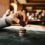 A hand placing a purple poker chip onto a stack on a dark gaming table in a blurred casino